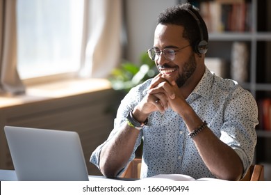Happy millennial african american man in glasses wearing headphones, enjoying watching educational webinar on laptop. Smiling young mixed race businessman holding video call with clients partners.